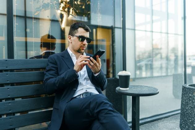 A businessman in a suit sits outdoors, using a smartphone while enjoying coffee. The modern urban setting highlights a blend of work and leisure. Sunglasses add sophistication Model Released PavelG_ID28721_848567_012 Copyright: xGERASIMENKOxPAVELx