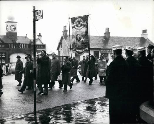 Demonstrationen 3. März 1961 - amerikanische Matrosen beobachten Demonstranten im Dunoon Submarine Depot Ship Proteus in Holy Loch. Anti-Atomkraft-Aktivisten waren in den Straßen von Dunoon - mit ihren Plakaten - Plakaten etc. - bald nach der Ankunft im Holy Loch des amerikanischen Polaris Submarine Depot Ship Proteus zu sehen... Foto zeigt: Matrosen der Prote s - beobachten Anti-Atomkraft-Aktivisten in den Straßen von Dunoon, Argyll - Fi th *** Demonstrations 3 March 1961 American Sailors monitor Demonstrators at Dunoon Submarine Depot Ship Proteus in Holy Hole Anti Nuclear power Activists Goods in the Roads from Dunoon with their Posters Posters ETC Soon to the Arrival at Holy Hole of American Polaris Submarine Depot Sh  Copyright: xUnitedxArchivesx xkpaxKeystonex 19610303_baf_k09_198