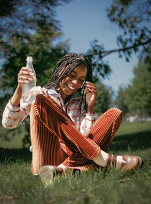 Creative Highlights Symbolbilder MR_ID29861_21585_EvynnJPendergrass.jpg A joyful African American woman with braided hair laughs heartily while sitting on grass and holding a water bottle. The sun shines brightly, adding warmth to the serene scene Model Released AnnaKapustina_ID29861_1008665_008 Copyright: xAnnaxKapustinax RECORD DATE NOT STATED