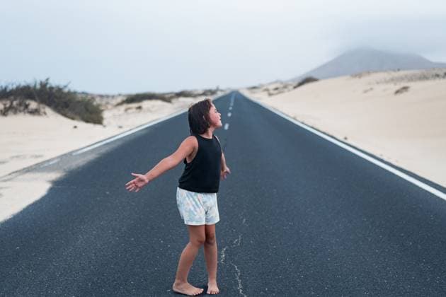 Creative Highlights Symbolbilder A child joyfully stands barefoot on an open road in Fuerteventura, Spain, surrounded by a serene desert landscape and distant mountains, under an overcast sky. Model Released HodeiUnzueta_ID29104_888711_011 Copyright: xHodeixUnzuetax