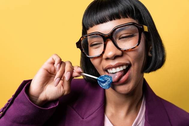 Creative Highlights Symbolbilder MR_ID30239_21746_OrianaDiaz.jpg Hispanic young woman with short hair and large glasses enjoying a blue heart-shaped lollipop. She s wearing a purple blazer and smiling against a vibrant yellow backdrop. Model Released ManuelRuiz_ID30239_1019449_007 Copyright: xManuelxRuizx RECORD DATE NOT STATED