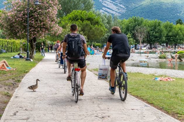 Nago-Torbole, Trentino, Italy - July 13, 2025: Holidaymakers cycle past bathing spots along the shore of Lake Garda near Torbole. The popular town is busy in summer, with tourists sharing cycle paths with walkers and animals *** Am Uferweg des Gardasees bei Torbole fahren Urlauber mit dem Fahrrad an Badestellen vorbei. Der beliebte Ort ist im Sommer gut besucht, Radwege teilen sich Touristen mit Spaziergängern und Tieren