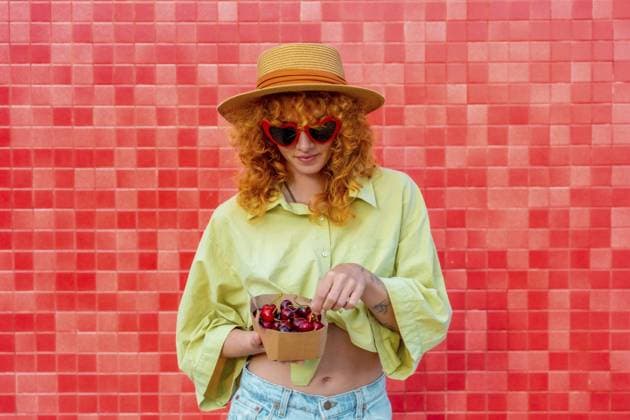 Modische rothaarige Frau mit herzförmiger Sonnenbrille und Strohhut, die frische Kirschen aus einer kleinen Pappschachtel vor einer leuchtend roten Kachelwand genießt Fashionable redhead woman with heart shaped sunglasses and straw hat enjoying fresh cherries from a small cardboard box against a vibrant red tiled wall Copyright: imageBROKER UnaixHuizi ibxuhu13859129.jpg ,model released, Symbolfoto ,property released
