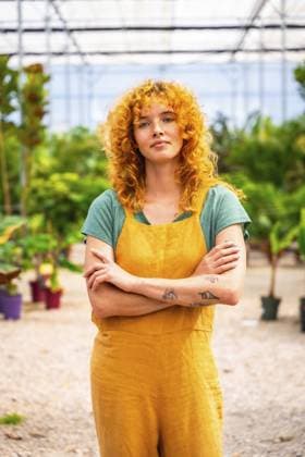 Junge Frau mit roten Locken und Tattoos, die einen gelben Overall trägt, steht mit verschränkten Armen in einem Gewächshaus und wirkt zuversichtlich Young woman with curly red hair and tattoos wearing yellow overalls, standing with crossed arms in plant nursery greenhouse, looking confident Copyright: imageBROKER UnaixHuizi ibxuhu14060281.jpg ,model released, Symbolfoto ,property released