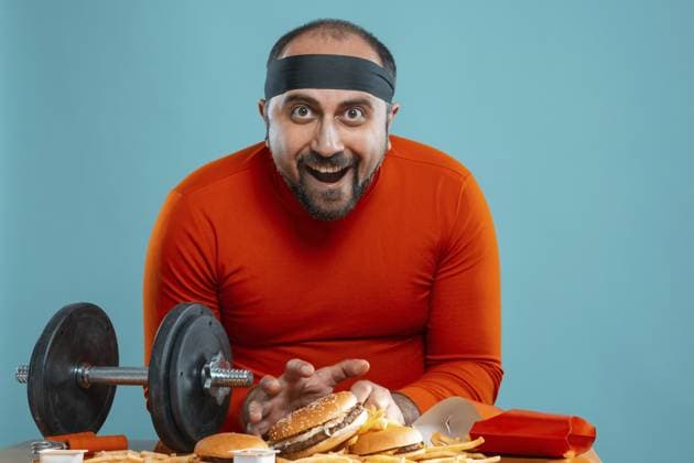 Close up Porträt eines brunet gut aussehend erwachsenen Mannes mit Bart, in einem roten Rollkragenpullover gekleidet, schwarzes Stirnband, Blick in die Kamera, während mit Hantel am Tisch mit Burger und Pommes frites gegen eine blaue Studio Hintergrund posiert. Aufrichtige Gefühle Konzept. Raum kopieren. Fast Food Close-up portrait of a brunet handsome adult man with beard, dressed in a red turtleneck, black headband, looking at the camera while posing with dumbbell at the table with burgers and french fries against a blue studio background. Sincere emotions concept. Copy space. Fast food Copyright: imageBROKER SerhiixNazarov ibxiqx14215998.jpg ,model released, Symbolfoto ,property released
