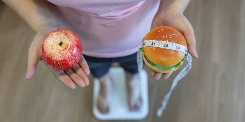 Eine Frau mit Übergröße steht auf einer Waage und wägt zwischen gesunder Ernährung und Genuss ab. In der einen Hand hält sie einen Apfel, in der anderen einen Burger A plus size woman stands on a scale, weighing her options between healthy eating and indulgence, holding an apple in one hand and a burger in the other Copyright: imageBROKER NATEExMEEPIAN ibxiqx14227147.jpg ,model released, Symbolfoto ,property released