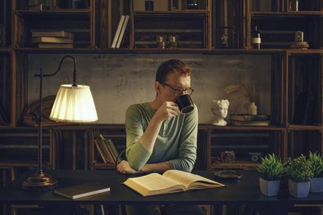 Ein Mann mittleren Alters sitzt an einem Schreibtisch in einem gemütlichen Zimmer und schlürft einen Kräutertee aus einer Tasse. Das sanfte Licht einer Lampe beleuchtet das Buch, das er vor dem Schlafengehen liest A middle-aged man sits at a desk in a cozy room, sipping a herbal tea from a mug. Soft light from a lamp illuminates the book he reads before going to bed Copyright: imageBROKER AlekseixIsachenko ibxfbm14380036.jpg ,model released, Symbolfoto ,property released