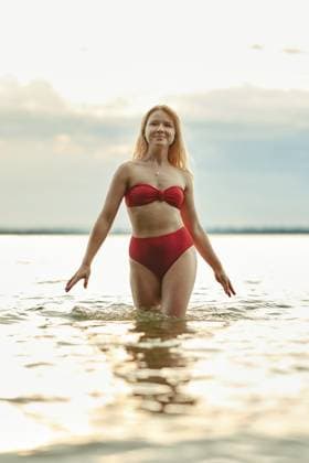 Eine junge Frau mit langen Haaren spaziert durch das seichte Wasser und präsentiert dabei einen leuchtend roten Badeanzug. Das weiche Licht des Abendhimmels schafft eine heitere Atmosphäre am Seeufer A young woman with long hair walks through shallow water, showcasing a vibrant red swimsuit. The soft light from the evening sky creates a serene atmosphere at a lakeside setting Copyright: imageBROKER AlekseixIsachenko ibxfbm14446180.jpg ,model released, Symbolfoto ,property released