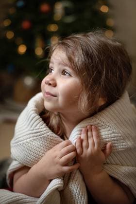 Child girl wrapped in sweater sitting near a decorated Christmas tree. Corby, England, United Kingdom R_JNDO2507302B-1795602-01 ,model released, Symbolfoto