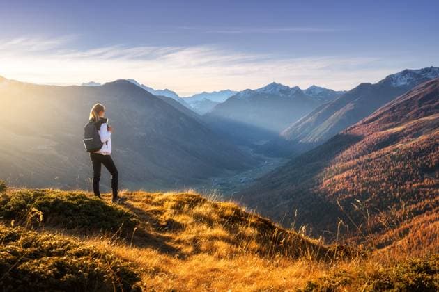 Young woman with backpack standing on grassy mountain ridge with view of valley with high mountain peaks during golden hour sunset in autumn. Hiking in Italy, Livigno. Girl in Alps in fall. Trekking Model Released DenisBilytskyi_ID29964_1012379_016 Copyright: xx
