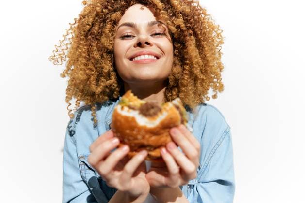 Happy young woman with curly hair enjoying a delicious burger against a bright white background, showcasing a vibrant and joyful food moment This vibrant image features a young woman with curly hair smiling widely while holding a juicy burger, set against a bright white background, representing happiness and indulgence in fast food, model released, , 96076012.jpg RECORD DATE NOT STATED