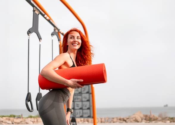 Smiling redhead sportswoman holding yoga mat at outdoor gym Smiling redhead sportswoman holding rolled yoga mat, looking away. Side view of fit, strong female athlete in sportswear standing at outdoor gym on a sunny day, model released, , 96351638.jpg RECORD DATE NOT STATED