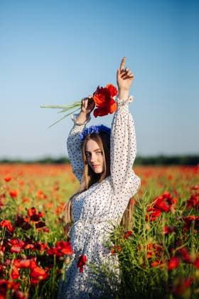 Beautiful young woman wearing a white dress and a flower crown is holding a bouquet of poppies in a poppy field Youthful female standing in blooming poppy meadow, wearing flowing white dress and floral headpiece, grasping vibrant red poppy bouquet, model released, , 64443222.jpg RECORD DATE NOT STATED