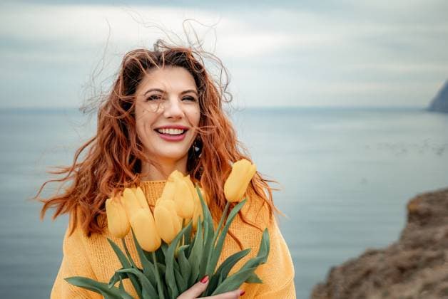 Portrait of a happy woman with hair flying in the wind against the backdrop of mountains and sea. Holding a bouquet of yellow tulips in her hands, wearing a yellow sweater Portrait of a happy woman with hair flying in the wind against the backdrop of mountains and sea. Holding a bouquet of yellow tulips in her hands, wearing a yellow sweater., model released, , 63322104.jpg RECORD DATE NOT STATED