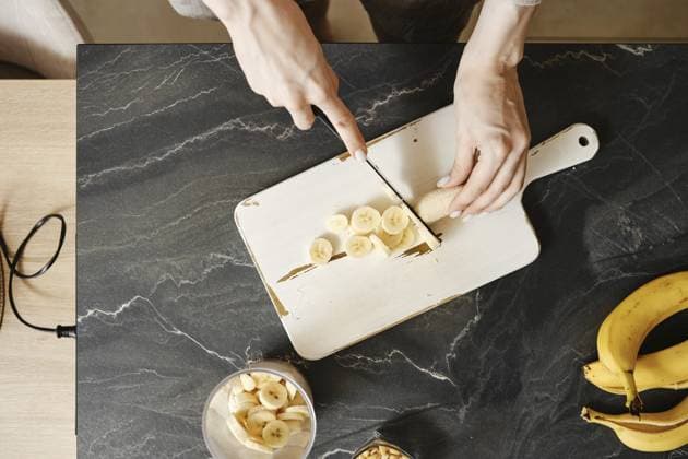Eine Person schneidet frische Bananen auf einem weißen Schneidebrett in einer gut beleuchteten Küche. Auf der Arbeitsplatte stehen eine Schale mit Bananen und ein Glasgefäß. Sonnenlicht strömt herein und schafft eine warme Atmosphäre A person slices fresh bananas on a white cutting board in a well-lit kitchen. The countertop features a bowl of bananas and a glass jar. Sunlight streams in, creating a warm atmosphere Copyright: imageBROKER AlekseixIsachenko ibxfbm14837568.jpg ,model released, Symbolfoto ,property released