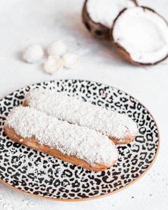 Eclairs with Shredded Coconut on Leopard Print Plate and Coconuts Two eclairs topped with shredded coconut rest on a leopard print plate, surrounded by halved coconuts and white confections on a white table., 64438240.jpg RECORD DATE NOT STATED