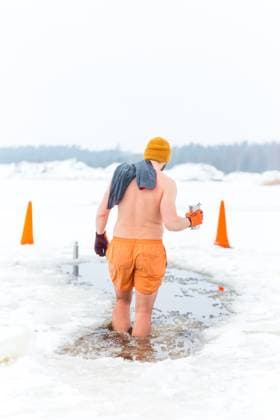 Person Preparing for Ice Swimming in a Snowy Rural Setting Individual in orange attire wades into a frozen body of water through a cut hole, surrounded by snow, traffic cones, and a forested winter backdrop., 64438686.jpg RECORD DATE NOT STATED