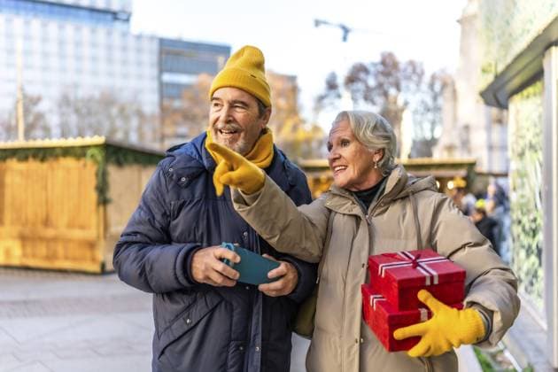 Fröhliches älteres Ehepaar mit Weihnachtsgeschenken in der Hand und einem Hinweis auf etwas Interessantes auf dem Weihnachtsmarkt in der Stadt Cheerful senior couple holding christmas gifts and pointing at something interesting at the christmas market in the city Copyright: imageBROKER UnaixHuizi ibxuhu14868978.jpg ,model released, Symbolfoto ,property released