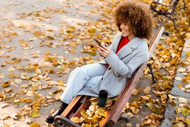 Enjoying a cozy moment in the park surrounded by autumn leaves while texting on a smartphone Sitting on a wooden bench in a park, a young woman smiles while texting on her smartphone, enjoying the vibrant autumn leaves that blanket the ground., model released, , 94965898.jpg RECORD DATE NOT STATED
