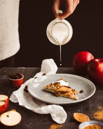 IMAGO Creative, Jahreszeit Herbst, Herbstanfang am 22. September Homemade apple galette with cream drizzle and fruit Cropped unrecognizable hand drizzling cream over homemade apple galette topped with cinnamon and raisins. Moody setting with red apples and gray background. Model Released VivianLyon_ID31138_1056277_005 Copyright: xVivianxLyonx RECORD DATE NOT STATED