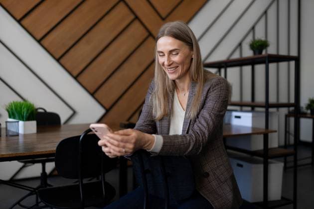 Professional woman smiling while using smartphone in modern office setting during daytime A woman with long hair and a blazer smiles while looking at her smartphone in a stylish office. Natural light highlights her relaxed demeanor among contemporary decor and furniture., model released, , 95412200.jpg RECORD DATE NOT STATED