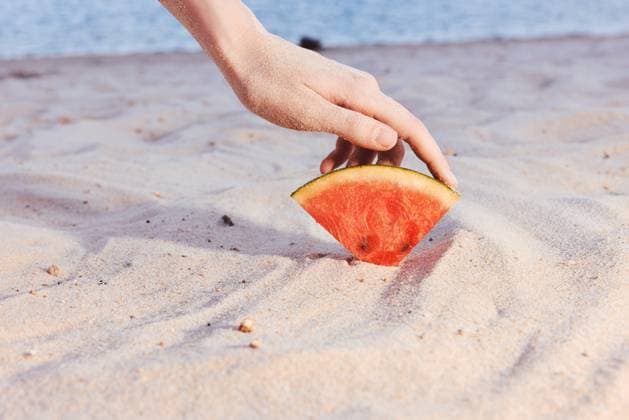 A hand placing a slice of watermelon on a sandy beach background, evoking summer and refreshment by the sea A hand gently places a slice of watermelon onto light colored sand at a beach, symbolizing summer, relaxation, and freshness, model released, , 96058526.jpg RECORD DATE NOT STATED