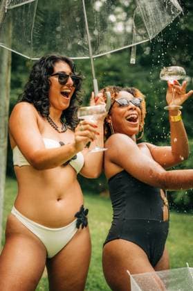 IMAGO Creative, Lifestyle, Feste im Garten #2 Two women in swimsuits laugh under umbrella with champagne splashes North Reading, Massachusetts, United States CR_FKNH250911-1827941-01 ,model released, Symbolfoto