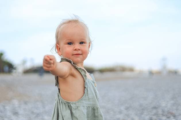 Creative Highlight Symbolbilder A one year old blonde girl playfully reaches her hand out on a pebble beach. Wearing a light dress, she smiles with a backdrop of a clear blue sky. Model Released AlinaKandybka_ID31292_1063416_012 Copyright: xAlinaxKandybkax RECORD DATE NOT STATED