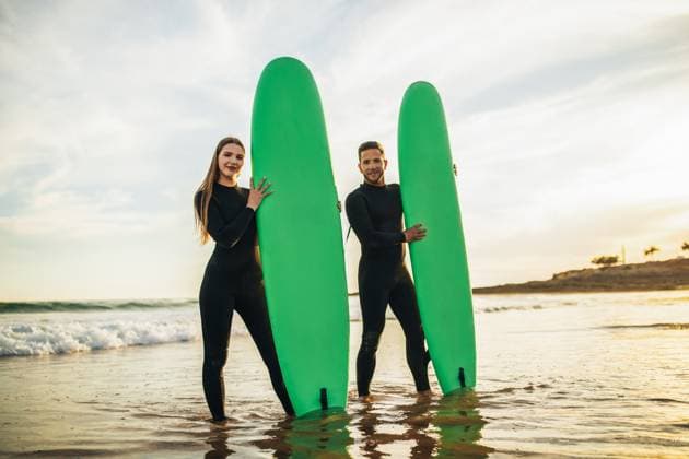 Couple Standing With Surfboards in Shallow Ocean Water at Sunset A couple in black wetsuits stands together in the shallow ocean water with their green surfboards. They are smiling and looking directly at the camera. The sun is setting behind them, model released, , 60769338.jpg RECORD DATE NOT STATED