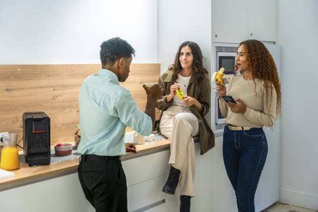 Verschiedene Kollegen plaudern und lächeln während einer zwanglosen Küchenpause im Büro, zwei Frauen halten Obst und ein Smartphone in der Hand, während ein männlicher Kollege spricht, entspannte Teamarbeit Diverse colleagues chatting and smiling during a casual office kitchen break, two women holding fruit and a smartphone while a male coworker talks, relaxed teamwork vibe Copyright: imageBROKER UnaixHuizi ibxuhu15197446.jpg ,model released, Symbolfoto ,property released