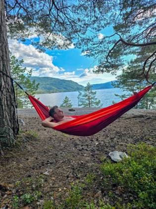 Entspannen in einer Hängematte an den ruhigen Gewässern Norwegens unter einem strahlend blauen Himmel A men enjoys a peaceful moment in a vibrant hammock, nestled between trees by Norways tranquil lake. The stunning landscape features lush greenery and distant mountains against bright skies. (License=RF) ,model released, Symbolfoto Copyright: xZoonar.com FokkexBaarssenx 23986871 ,model released, Symbolfoto ,property released