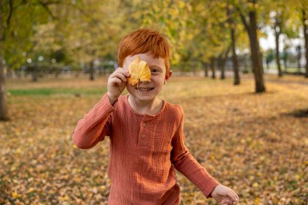 Red-haired child smiling outdoors covering eye with yellow autumn leaf model released, Symbolfoto, MBLF01042
