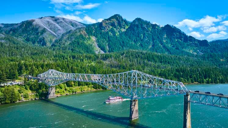 Aerial of Bridge of the Gods Over River with Paddle Steamer in Forested Mountains Aerial view of the Bridge of the Gods spanning the Columbia River in Cascade Locks, with a historic paddle steamer cruising below, set against a scenic backdrop of lush, tree-covered mountains in the Pacific Northwest., 60921582.jpg RECORD DATE NOT STATED