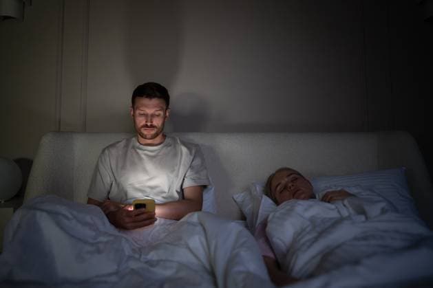 Müder Mann benutzt nachts im Bett liegend sein Smartphone, während seine Frau neben ihm im dunklen Schlafzimmer schläft Exhausted man awake alone in bed scrolling phone due to insomnia and digital addiction on late hours. Sleepless male with social media dependency, difficulty falling asleep, partner ignoring spouse (License=RF) ,model released, Symbolfoto Copyright: xZoonar.com DmitriixMarchenkox 24118661 ,model released, Symbolfoto ,property released