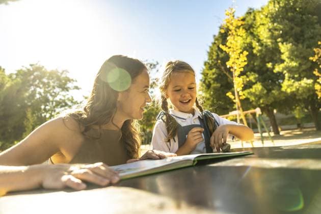 Mutter und Tochter lächeln und lachen, während sie gemeinsam an einem Tisch im Freien in einem sonnigen Park oder auf einem Schulhof ein Buch lesen und dabei warme Zeit und Lernen genießen Mother and daughter smiling and laughing while reading a book together at an outdoor table in a sunny park or school playground, enjoying warm quality time and learning Copyright: imageBROKER UnaixHuizi ibxuhu15439999.jpg ,model released, Symbolfoto ,property released