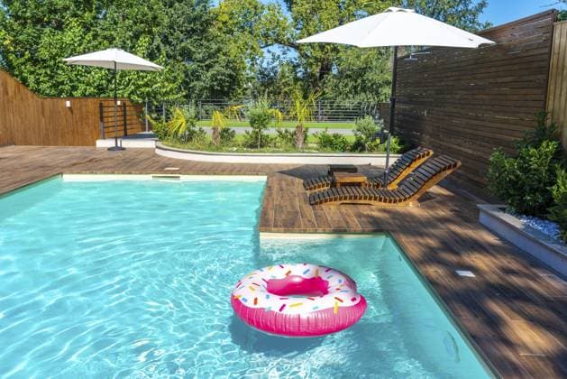 Hinterhofpool mit türkisfarbenem Wasser, Holzdeck, Sonnenschirmen, Liegestühlen und rosafarbenem Donut Float Backyard swimming pool with turquoise water, wooden deck, umbrellas, lounge chairs, and pink donut float Copyright: imageBROKER MariaxKraynova ibxiqx15511087.jpg