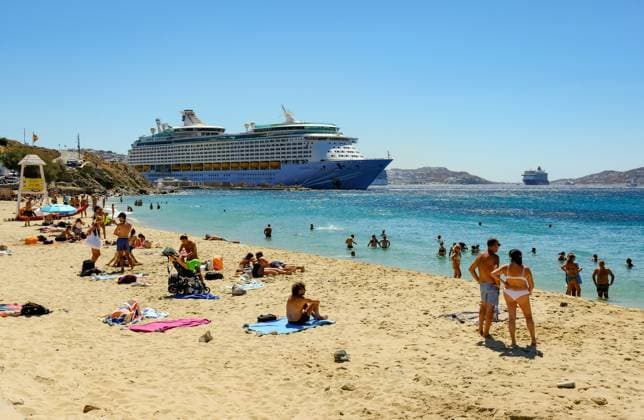 Mykonos, Kykladen, Griechenland - Kreuzfahrtschiff Voyager of the Seas liegt in der Hafenbucht Mykonos. Vorne baden Touristen und Einheimische am Sandstrand Paralia Agios Stefanos. Mykonos Mykonos Griechenland *** Mykonos, Cyclades, Greece Cruise ship Voyager of the Seas moored in Mykonos harbor Bay In front, tourists and locals bathe on the sandy beach Paralia Agios Stefanos Mykonos Mykonos Greece