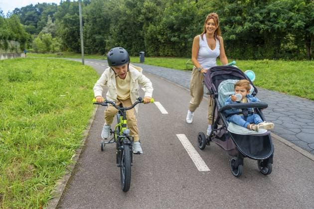 Mutter geht mit ihrem Baby im Kinderwagen spazieren, während der ältere Sohn auf einem gepflasterten Weg Fahrrad fährt und die gemeinsame Zeit im Freien genießt, umgeben von Natur und Sonnenlicht Mother walking with her baby in a stroller while older son riding his bicycle on a paved path, enjoying quality time together outdoors, surrounded by nature and sunlight Copyright: imageBROKER UnaixHuizi ibxuhu15661025.jpg ,model released, Symbolfoto ,property released