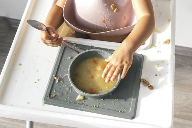 Das Baby lernt, selbständig zu essen, sitzt in einem Hochstuhl mit einer Schüssel Suppe, macht Unordnung, während es das Essen anfasst und einen Löffel hält, trägt ein Silikon Lätzchen Baby learning to eat independently, sitting in a high chair with a bowl of soup, making a mess while touching the food and holding a spoon, wearing a silicone bib Copyright: imageBROKER EvgeniiaxGordeeva ibxiqx15555351.jpg ,model released, Symbolfoto ,property released