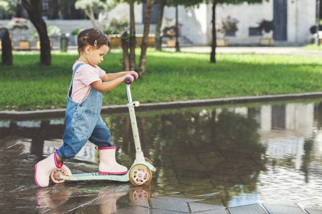 Ein kleines Mädchen in Jeansoveralls und pinken Gummistiefeln fährt mit einem Roller durch eine Pfütze in einem Park, konzentriert und entschlossen, während sie durch das Wasser planscht A little girl in denim overalls and pink rubber boots rides a scooter through a puddle in a park, focused and determined as she splashes through the water Copyright: imageBROKER EvgeniiaxGordeeva ibxiqx15555390.jpg ,model released, Symbolfoto ,property released