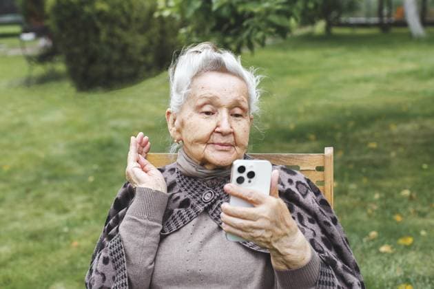 Ältere Frau sitzt draußen auf einem Stuhl und winkt während eines Videoanrufs auf dem Smartphone, Konzept des Alterns und der Technologie Elderly woman sitting on chair outdoors and waving during video call on smartphone, concept of aging and technology Copyright: imageBROKER EvgeniiaxGordeeva ibxiqx15556008.jpg ,model released, Symbolfoto ,property released