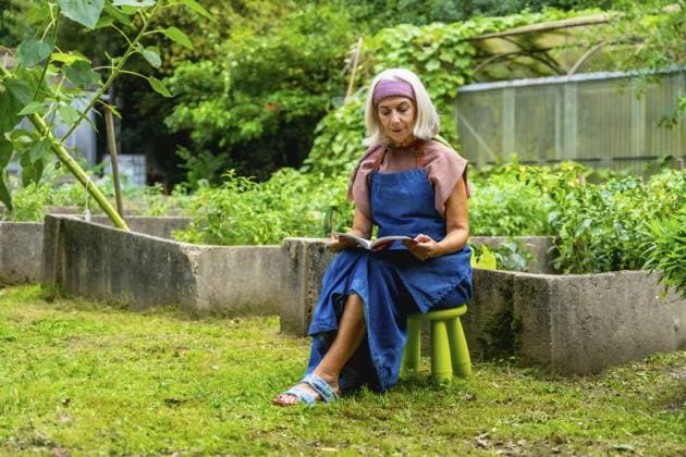 Seniorin mit Schürze und Stirnband genießt einen ruhigen Moment beim Lesen eines Buches, während sie auf einem Hocker in ihrem grünen Garten neben Hochbeeten sitzt Senior woman wearing an apron and headband, enjoying a quiet moment reading a book while sitting on a stool in her green outdoor garden next to raised beds Copyright: imageBROKER UnaixHuizi ibxuhu15681129.jpg ,model released, Symbolfoto ,property released