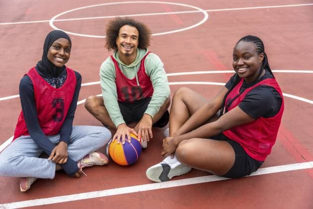 Verschiedene junge Freunde in Sporttrikots und Hijab, die lächeln, während sie zusammen mit einem Ball auf einem Basketballplatz im Freien sitzen, was Teamwork und aktive Jugend repräsentiert Diverse young friends wearing sport jerseys and a hijab, smiling while sitting together on an outdoor basketball court with a ball, representing teamwork and active youth Copyright: imageBROKER UnaixHuizi ibxuhu15750160.jpg ,model released, Symbolfoto ,property released