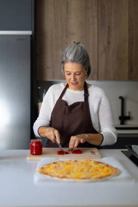 Senior woman preparing homemade pizza topping in kitchen Senior woman cutting red paprika on a wooden board, preparing fresh topping for a homemade pizza on a kitchen counter Model Released SloomStudio_ID32523_1108175_009 Copyright: xcristinaderojasfotografiax