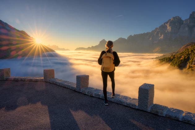 Woman with yellow backpack stands on a viewpoint above a sea of clouds at sunset in Swiss Alps. Girl, sun rays illuminate the alpine mountain peaks and misty valley. Travel in Switzerland. Hiker Model Released DenisBilytskyi_ID32879_1119722_019 Copyright: xx