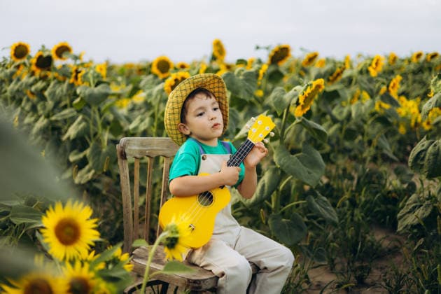 Child exploring a sunflower field with joy A joyful child plays a ukulele while sitting in a vibrant sunflower field. The sunny day and flourishing flowers create a delightful scene, capturing the essence of childhood wonder. Model Released Ptashkimenko_ID32371_1103894_003 Copyright: xx RECORD DATE NOT STATED