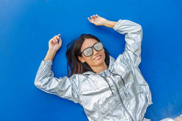 An Asian woman wearing a sparkling silver suit lies on a blue background, smiling confidently. Her rhinestone studded sunglasses add a touch of glamour and style. Model Released JuanAlgar_ID31575_1073643_014 Copyright: xJuanxAlgarx RECORD DATE NOT STATED