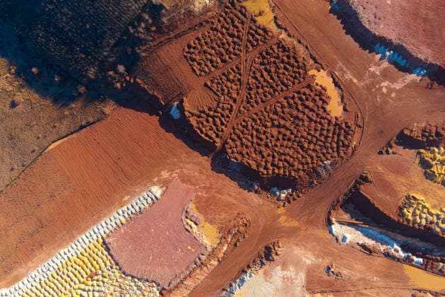 Luftaufnahme eines industriellen Steinbruchs von oben mit roten und gelben Erdhaufen, die in Reihen angeordnet sind und eine Abbaustufe zeigen. Abstract drone shot of an open-pit mine featuring organized mounds of red clay and bright yellow mineral deposits. The textured earth piles and muddy pools highlight industrial extraction. (License=RF) Copyright: xZoonar.com AndriixChagovetsx 24340352