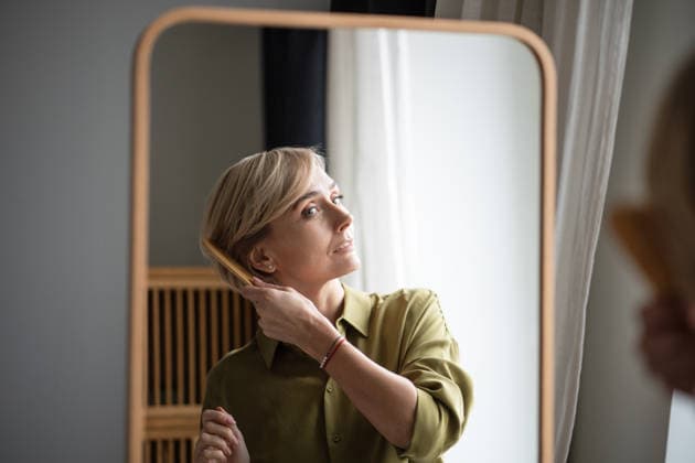 Eine positive Frau mittleren Alters genießt ihren neuen Haarschnitt und kämmt ihr Haar mit einem Holzkamm vor einem Spiegel. Attractive middle aged woman enjoying new haircut, standing in front of mirror in room and combing hair with wooden comb, fixing hairstyle and outlook in reflection with satisfied positive expression. (License=RF) ,model released, Symbolfoto Copyright: xZoonar.com DmitriixMarchenkox 24399949 ,model released, Symbolfoto ,property released