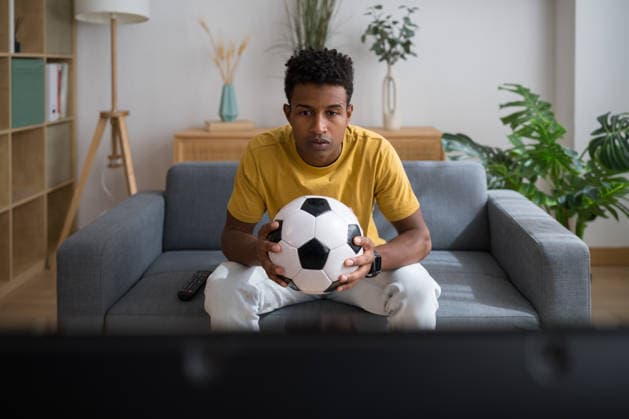 Man holding soccer ball focused watching sport on sofa indoors model released, Symbolfoto property released, ATSF00086